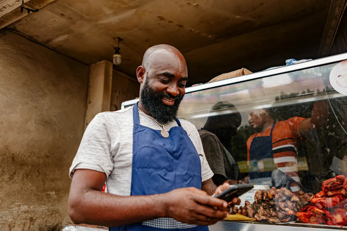 A small-business owner at his food stall smiling at his mobile phone.