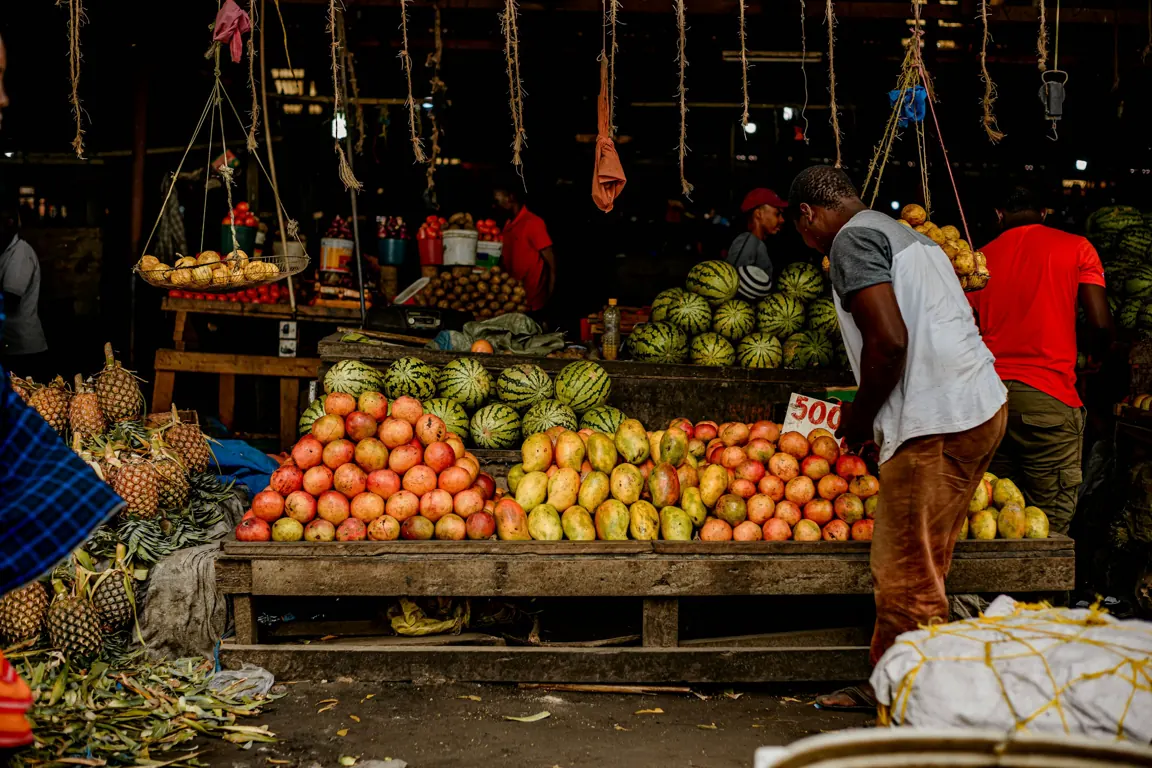 A market vendor standing behind a fruit stall in a covered market.
